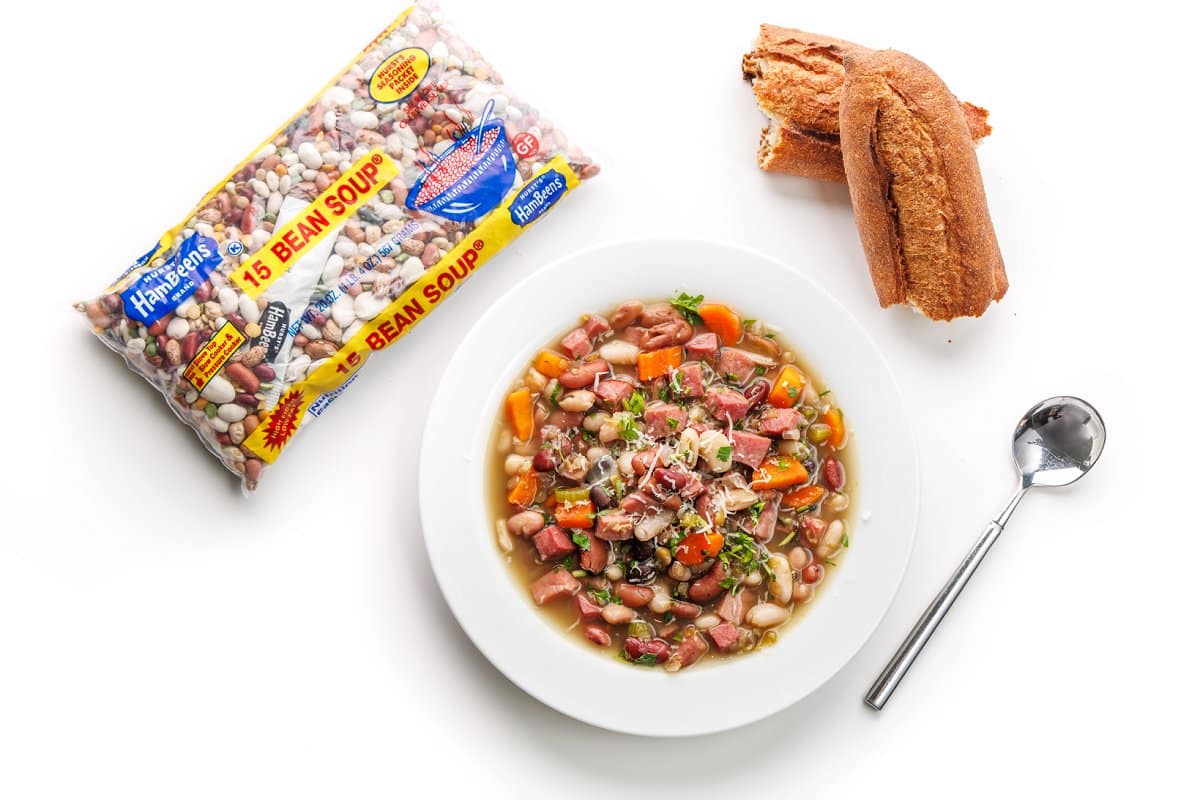 15 bean soup with ham in a white bowl with a silver spoon next to a torn loaf of bread and a Hurst's 15 bean soup package.