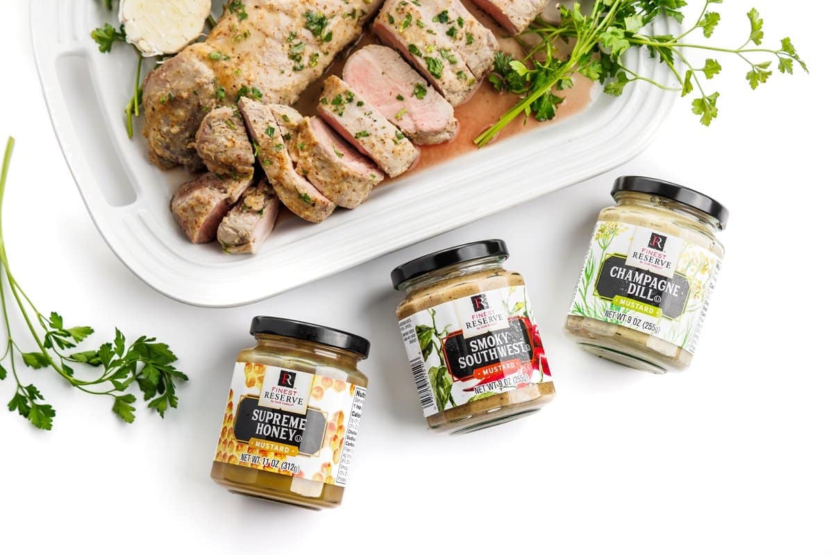 Cooked pork tenderloin on a platter next to containers of Finest Reserve mustard.