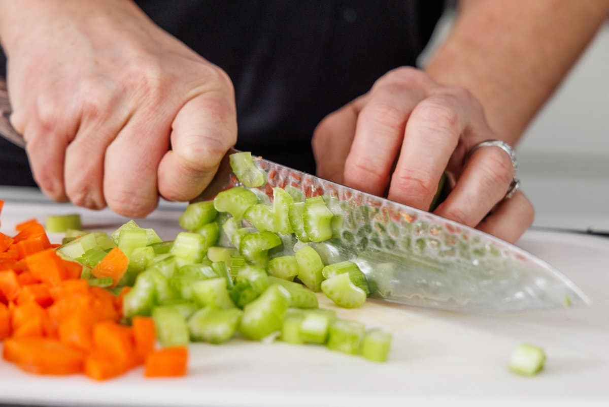 Liz dicing celery and carrot on a cutting board with a large, sharp knife.