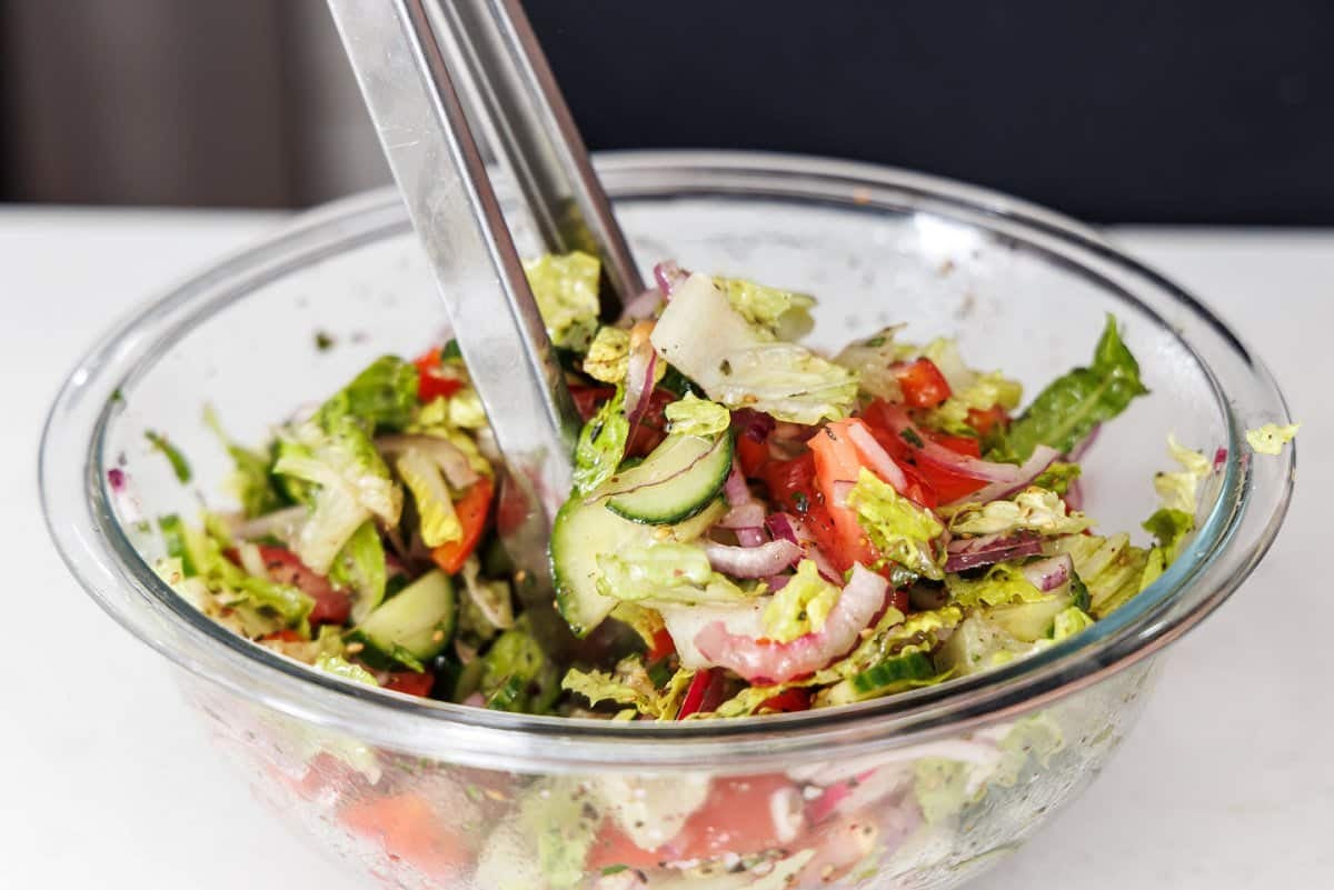 Tossing together a Lebanese salad in a glass bowl with metal tongs.