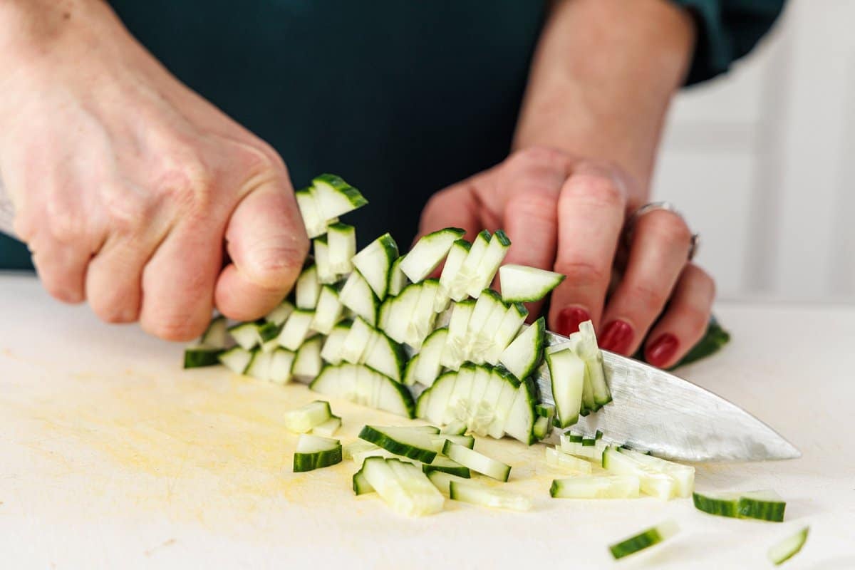 LIz dicing a cucumber on a cutting board with a large knife.
