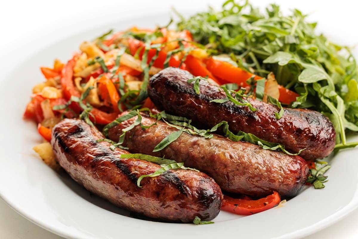 Close up of 3 grilled bratwursts on a white plate, with arugula and grilled veggies behind them on the plate.