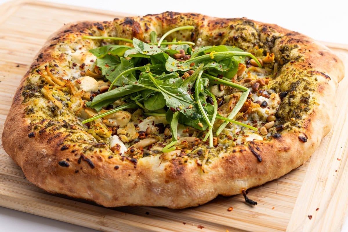 Cooked pesto chicken pizza with goat cheese and pine nuts on a cutting board.