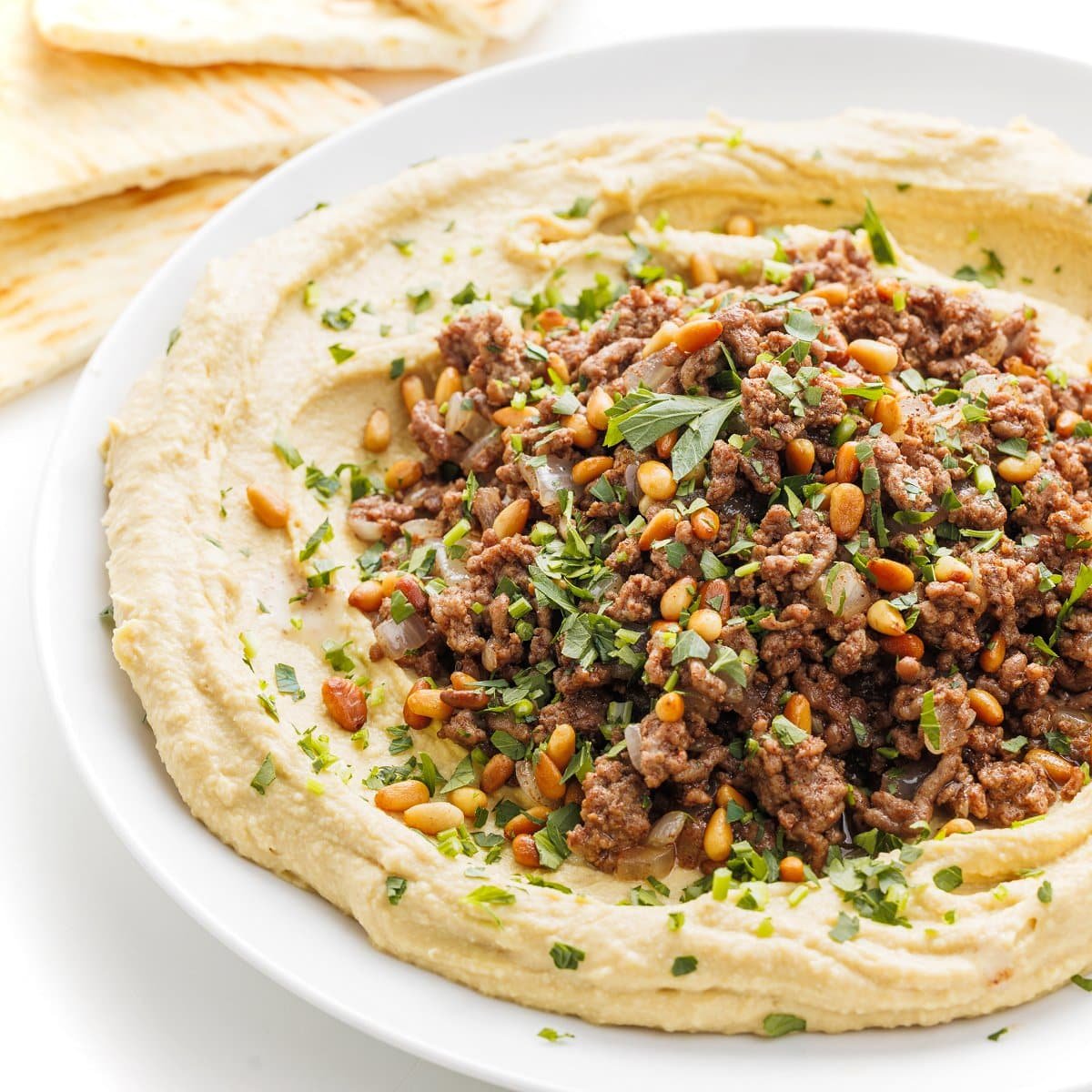 Hummus with ground lamb and toasted pine nuts spread on a white plate.