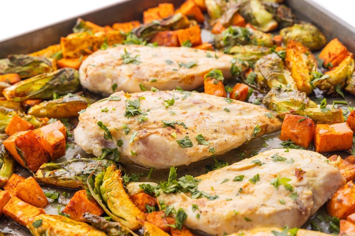 Three chicken breasts garnished with parsley on a sheet pan with roasted fall veggies.