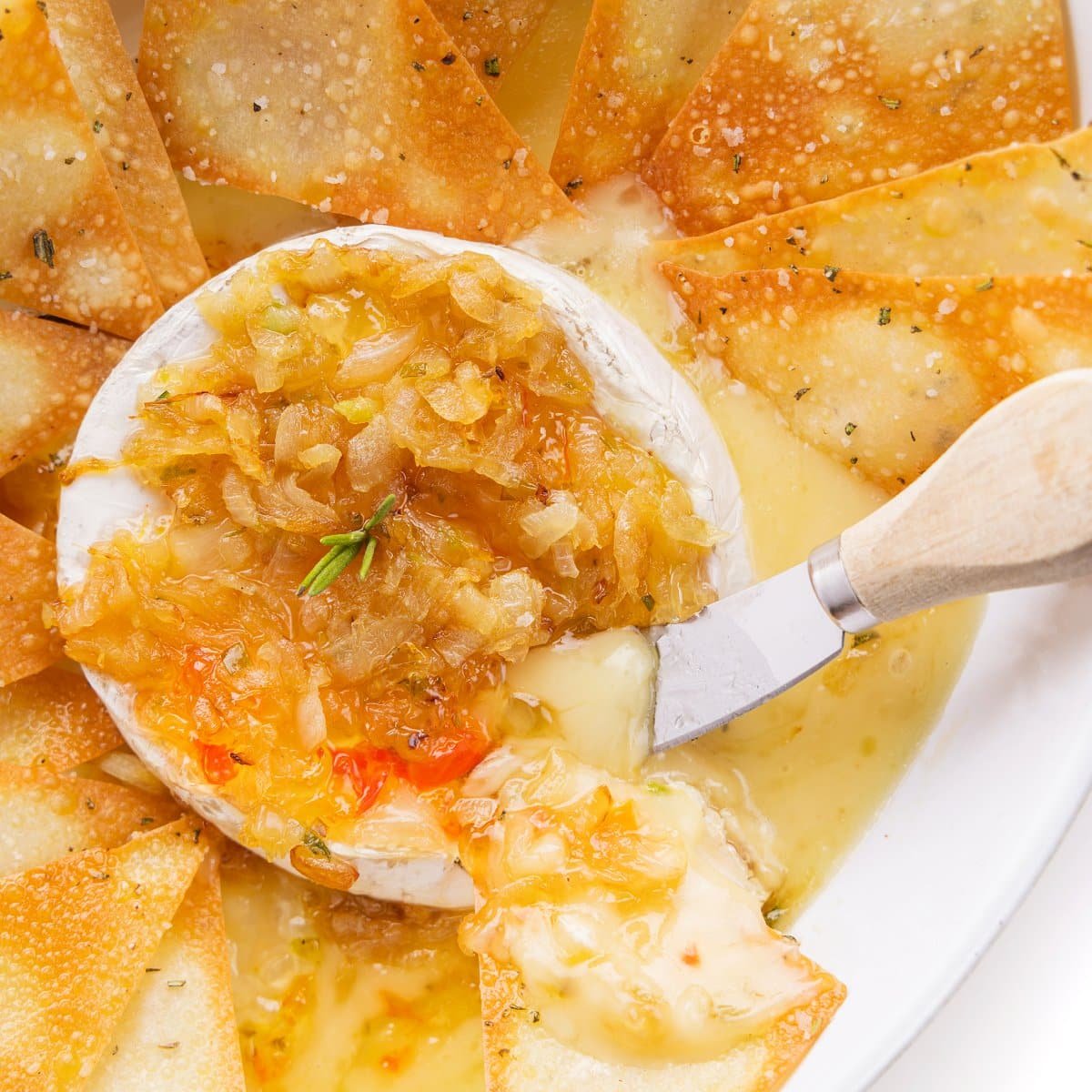 Wheel of baked brie with caramelized onions with a cheese knife and wonton crackers.