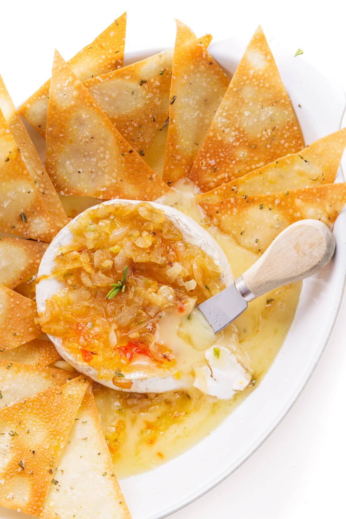 A cheese knife in a wheel of baked brie with caramelized onions in a baking dish with wonton crackers.