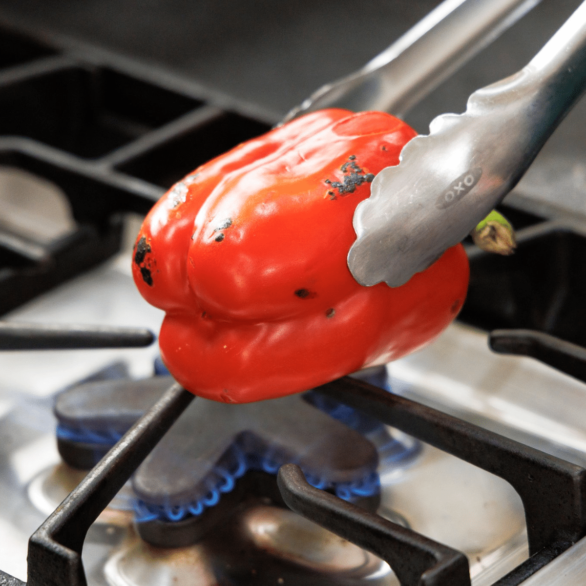 Tongs holding red bell pepper over gas burning stove flame.