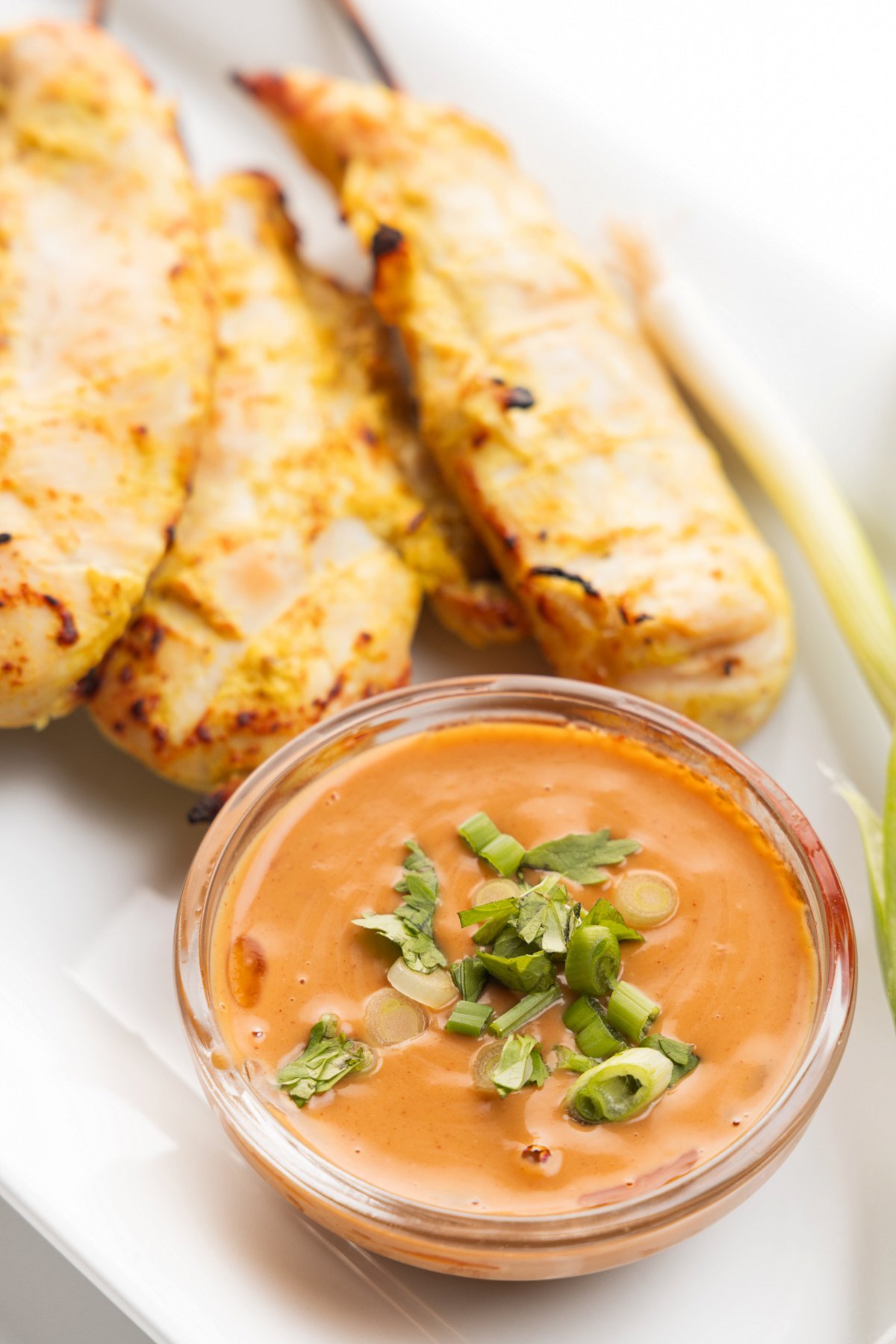 Small glass bowl of peanut sauce topped with fresh herbs next to chicken satay skewers.